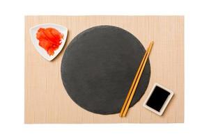 Emptyround black slate plate with chopsticks for sushi, ginger and soy sauce on brown sushi mat background. Top view with copy space for you design photo