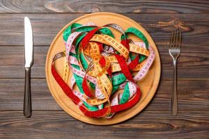 Heap of colorful measuring tape instead of spaghetti in round plate on wooden background. Top view of healthy eating concept photo