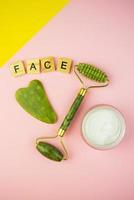 herramientas de masaje facial verde gua sha. rodillo de jade cuarzo verde sobre fondo rosa-amarillo. tarro de crema, letras de cara en letras de madera. foto