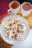Two cups of black tea stand on a wooden tray. Fresh and fragrant cinnamon rolls close up lie on a plate with polka dots, beautiful morning. Close-up. Romantic morning. View from above