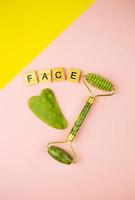 herramientas de masaje facial verde gua sha. rodillo de jade cuarzo verde sobre fondo rosa-amarillo. la cara de inscripción en letras de madera. foto