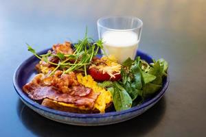 desayuno muy brillante y saludable en un plato: tocino, huevo, ensalada, tomate, queso, un vaso de yogur. foto