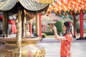 mujer asiática con vestido tradicional cheongsam qipao rezando con varitas de incienso por la mejor bendición y buena suerte en el templo budista chino. celebrar el año nuevo lunar chino. foto