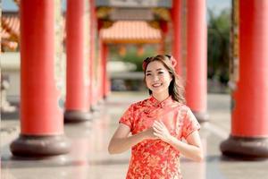 mujer asiática con vestido tradicional cheongsam qipao con gesto de felicitación en el templo budista chino. sonrisa de emoción. concepto de año nuevo en cadena. emoción sonrisa foto