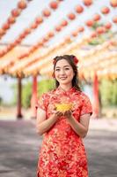 Vertical picture. A young asian woman wearing traditional cheongsam qipao dress holding ancient gold money in Chinese Buddhist temple. Celebrate Chinese lunar new year, festive season holiday. photo