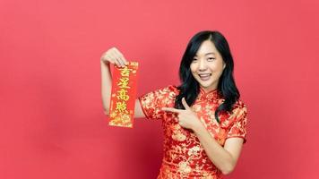 Young woman wearing traditional cheongsam qipao  costume holding and pointing blessing fortune card isolated on red background. Chinese text means great luck great profit photo
