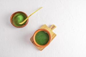 Top view of freshly made matcha green tea in a ceramic cup and matcha powder in a wooden bowl with a bamboo measuring spoon. white background.