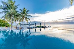 Tropical beach resort with lounge chairs and umbrellas and coco palm trees. Beautiful calm morning, infinity pool close to sea and beach, sky reflection. Luxury peaceful tropical vacation