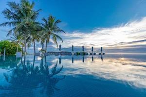 Tropical beach resort with lounge chairs and umbrellas and coco palm trees. Beautiful calm morning, infinity pool close to sea and beach, sky reflection. Luxury peaceful tropical vacation photo