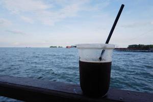 enfoque selectivo en una taza de plástico que contiene café helado negro con vistas al mar en el fondo foto