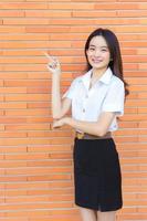 Portrait of an adult Thai student in university student uniform. Asian beautiful girl standing to present something confidently on brick walls background. photo