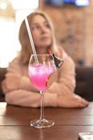 Fruity pink cocktail with a slice of lime on a table in a cafe. In the background, a blurry figure of a woman sitting at a table photo