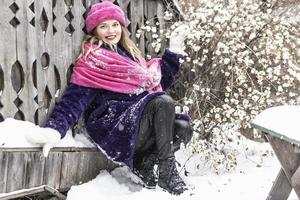 Portrait of a smiling young woman in a bright pink scarf and hat, white mittens and a purple fur coat outdoors in a city park in winter. photo