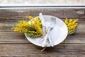 Fork knife with plate and mimosa flowers on wooden table on window background. spring Easter holiday dinner. photo