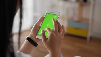 Closeup vertical of hand woman using smartphone with green screen while sitting in living room. Blank digital smartphone in hand girl. Showing content videos blogs tapping on center screen.