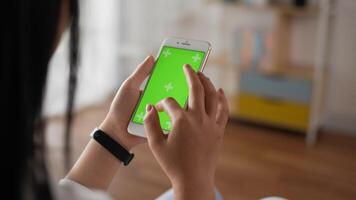 primer plano vertical de la mujer de la mano usando un teléfono inteligente con pantalla verde mientras está sentado en la sala de estar. teléfono inteligente digital en blanco en la mano chica. mostrando blogs de videos de contenido tocando en el centro de la pantalla.