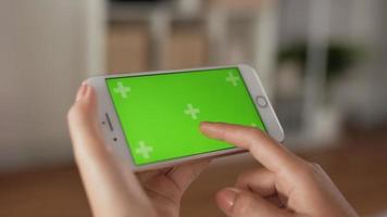 Closeup horizontal of hand woman using smartphone with green screen while sitting in living room. Blank digital smartphone in hand girl. Showing content videos blogs tapping on center screen.