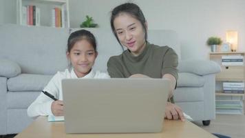 lección de enseñanza de madre para hija por computadora portátil. una niña asiática aprende en casa. hacer la tarea con la amable ayuda de la madre, animar para el examen. niña de asia feliz educación en el hogar. mamá aconseja educación juntos. video