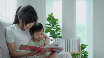 glückliche asiatische mutter entspannen und lesen buch mit babyzeit zusammen zu hause. eltern sitzen mit tochter auf dem sofa und lesen eine geschichte. Lernentwicklung, Kinderbetreuung, Lachen, Bildung, Geschichtenerzählen, Üben. video