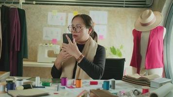 Asian middle-aged female fashion designer works in studio by talking on mobile phone about ideas and drawing sketches for dress design collection orders. Professional boutique tailor SME entrepreneur. video