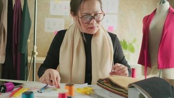 asiatische modedesignerin mittleren alters arbeitet im studio, schneidet und wählt stoff- und fadenfarben mit zeichnungsskizzen für kleiderdesign-kollektionen. professioneller Boutique-Schneider-Unternehmer. video