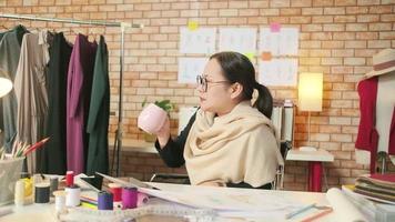 Asian middle-aged female fashion designer works in creative studio, sketching, drinking coffee, thinking and crushing ideas in dress design collections, and professional boutique tailor entrepreneur. video