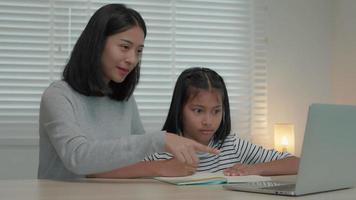 lección de enseñanza de madre para hija por computadora portátil. una niña asiática aprende en casa. hacer la tarea con la amable ayuda de la madre, animar para el examen. niña de asia feliz educación en el hogar. mamá aconseja educación juntos. video