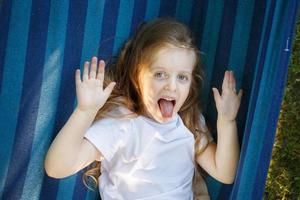 portrait of a little cute girl with long hair resting on a hammock in the garden and showing tongue photo