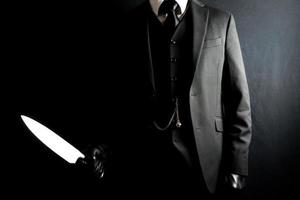 retrato de hombre con traje oscuro y guantes de cuero sosteniendo un cuchillo afilado sobre fondo negro. asesino de caballeros bien vestido.