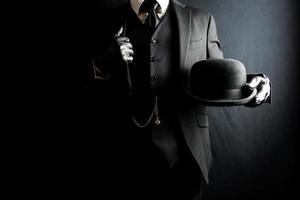 Portrait of Man in Dark Suit and Leather Gloves Holding Bowler Hat on Black Background. Concept of Classic British Gentleman photo