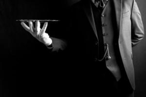retrato de mayordomo o camarero con traje oscuro y guantes blancos sosteniendo expertamente una bandeja de plata sobre fondo negro. concepto de industria de servicios y hospitalidad profesional. foto