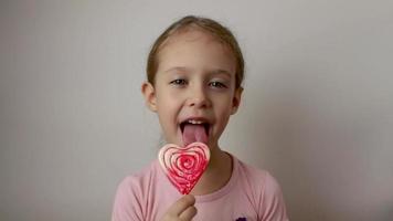 una linda niña lame una piruleta grande en forma de corazón rojo y blanco. fondo blanco aislado video