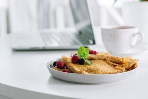 desayuno delicioso. mesa de escritorio blanca con computadora portátil y crepes frescos, taza de té. concepto de autónomo. desayuno saludable. foto