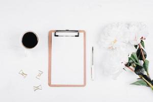 Workplace with cup of coffee, flowers peonies, pen, clipboard. Flat lay, top view, mockup photo