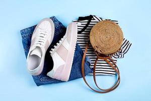 Blue jeans shorts, sneakers, striped shirt and rattan bag on blue background. Flat lay, top view photo