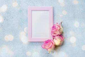 Holiday card concept. Photo frame with place for text and pink roses on blue background. Top view, flat lay. Mock up
