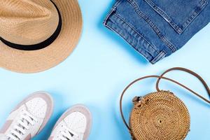 Blue jeans shorts, sneakers, hat and modern rattan bag on blue background. Flat lay, top view. Summer outfit concept photo