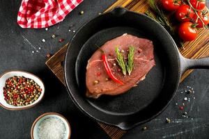 Raw meat in black cast-iron pan. Pork steaks with spices, herbs dark background. Top view