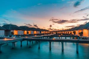 Beautiful sky and sea water, luxury tropical resort, water villas with lights under sunset. Summer island background, luxurious vacation banner. Tranquil, relaxing mood, inspirational nature landscape photo