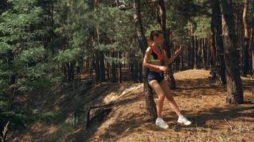 una chica atlética linda y sonriente se para cerca de un árbol en el bosque y sostiene el teléfono en la mano video