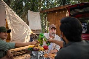 grupo de turistas amigos acampando y haciendo una barbacoa y bebiendo cerveza-alcohol y rompiendo una botella de cerveza con la fiesta junto con disfrute y felicidad en verano foto