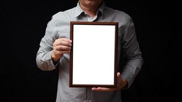 A blank diploma or a mockup certificate in the hand of a man employee wearing shirt on black background. The vertical picture frame is empty and the copy space. photo