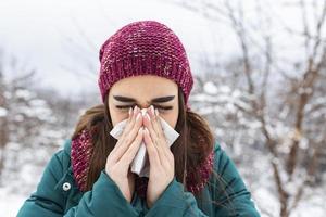 mujer joven enferma y estornuda en papel de seda. niña sonarse la nariz al aire libre. coger frío en invierno. mujer triste y molesta con mala expresión estornuda y tiene secreción nasal foto