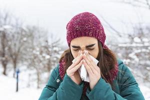 Young woman sick and sneeze into tissue paper. Girl blowing nose outdoors.Catching cold in winter. Upset gloomy woman with ill expression sneezes and has running nose