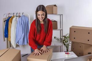 Young woman, owner of small business packing product in boxes, preparing it for delivery. Women packing package with her products that she selling online. Beautiful woman moving to another place photo