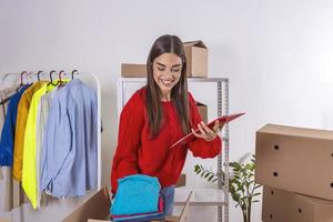 Young woman holding clipboard,looking at laptop for adress of customer. Small business owner,selling online,packing boxes and delivery photo