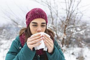 Young woman suffering from a seasonal cold and flu blowing her nose on a handkerchief as she stands outdoors on winter. Healthcare and medical concept photo