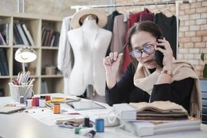 Asian middle-aged female fashion designer works in studio by talking on mobile phone about ideas and drawing sketches for dress design collection orders. Professional boutique tailor SME entrepreneur. photo