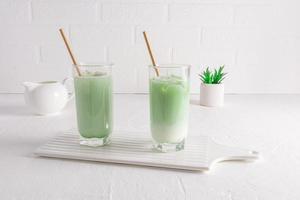 a healthy matcha latte drink with milk in tall glasses with straw on a white ceramic board against a white wall. natural organic drink.