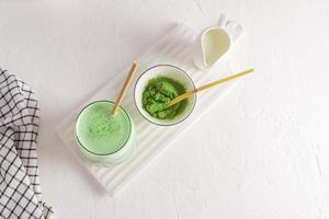 Top view of a white ceramic board with a glass of green match latte, tea powder and a jug of milk. freshly prepared drink. a health remedy. photo